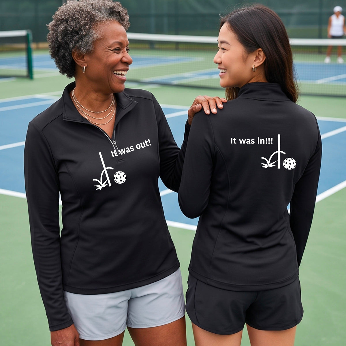 Two women wearing a black  quarter-zip shirt with a white design of ‘IT WAS OUT!’ and a colorful pickleball on the front, and the back design with a white ‘IT WAS IN!!!’ and a colorful pickleball, standing on a pickleball court.