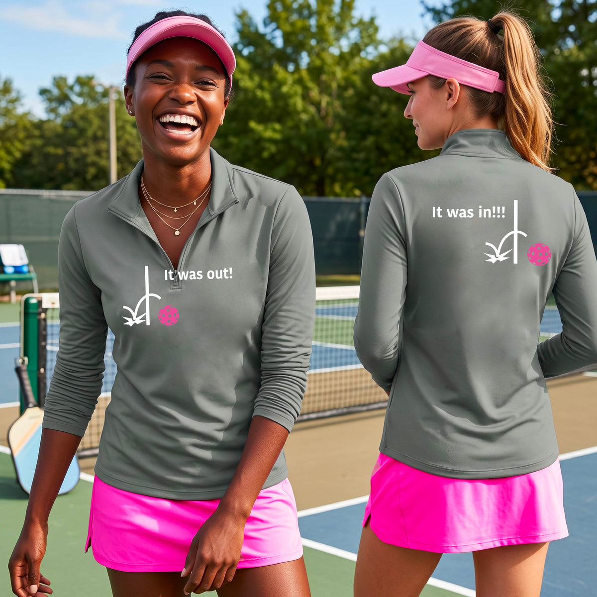 Two women wearing a gray quarter-zip shirt with a white design of ‘IT WAS OUT!’ and a colorful pickleball on the front, and the back design with a white ‘IT WAS IN!!!’ and a colorful pickleball, standing on a pickleball court.