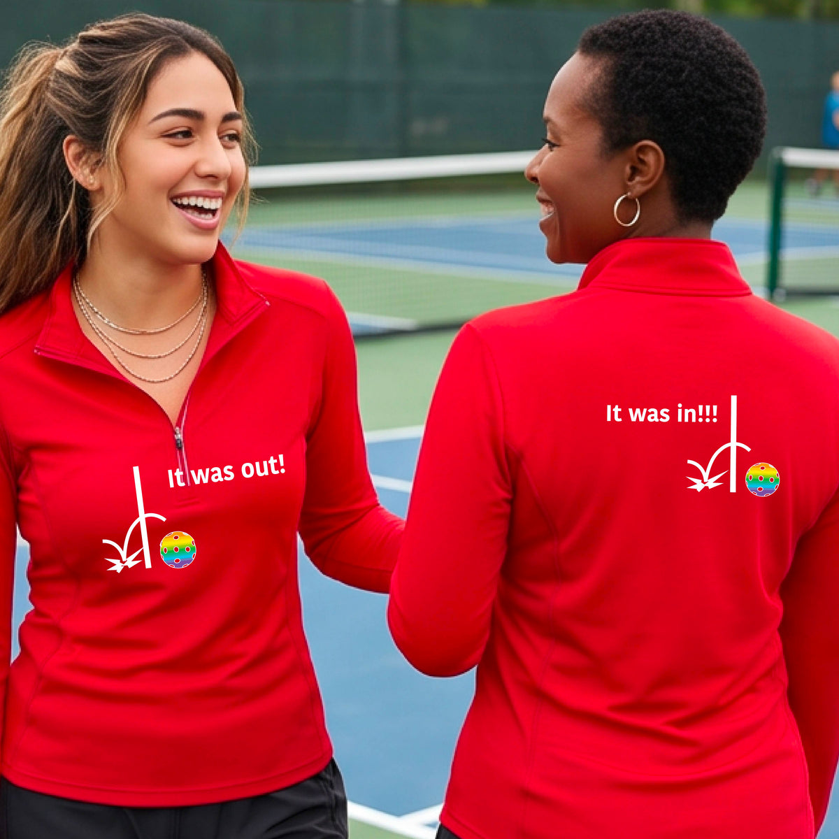 Two women wearing a red quarter-zip shirt with a white design of ‘IT WAS OUT!’ and a colorful pickleball on the front, and the back design with a white ‘IT WAS IN!!!’ and a colorful pickleball, standing on a pickleball court.