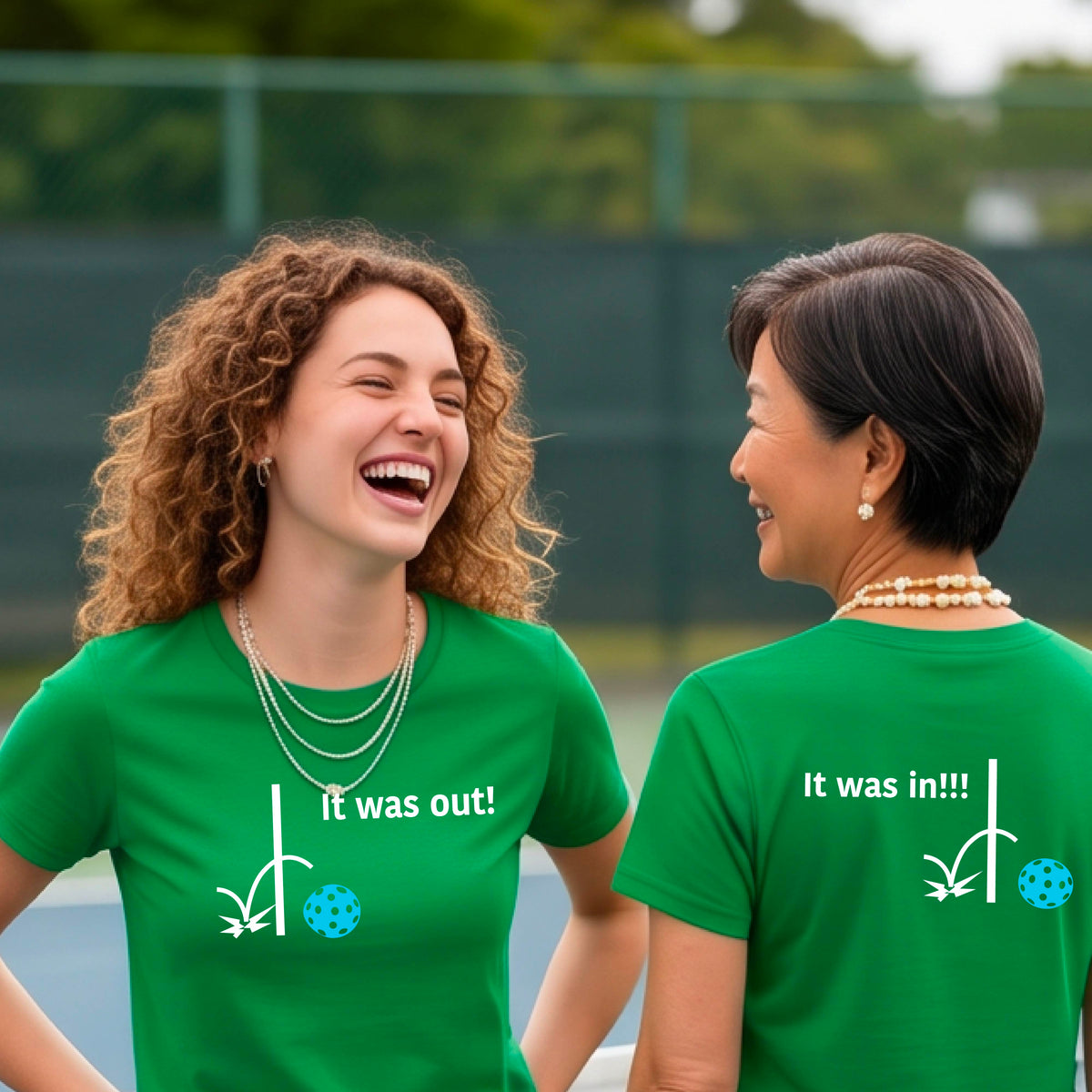 Two women showing a green short-sleeve t-shirt with a white design of ‘IT WAS OUT!’ and a colorful pickleball on the front, and the back design with a white ‘IT WAS IN!!!’ and a colorful pickleball, standing on a pickleball court.
