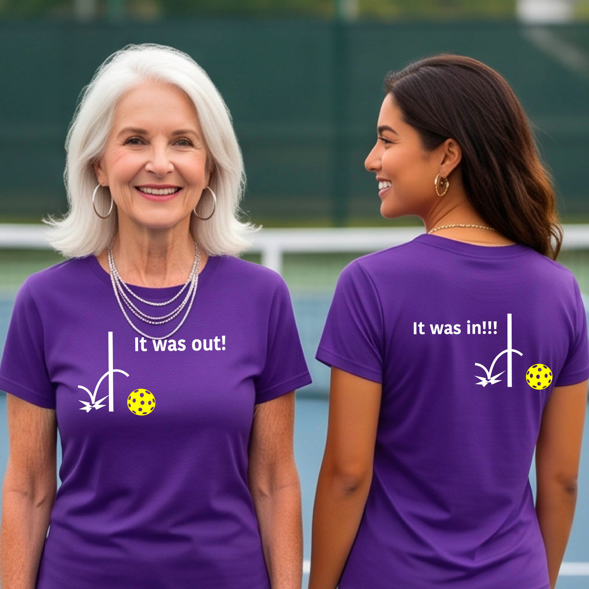 Two women showing a purple short-sleeve t-shirt with a white design of ‘IT WAS OUT!’ and a colorful pickleball on the front, and the back design with a white ‘IT WAS IN!!!’ and a colorful pickleball, standing on a pickleball court.