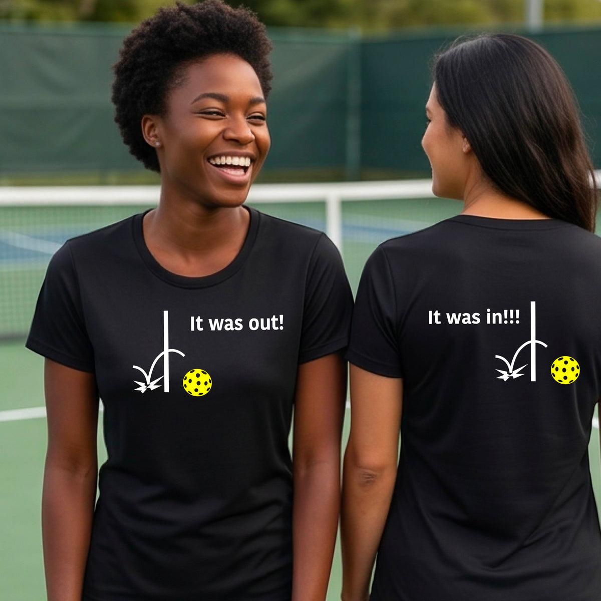 Two women showing a black short-sleeve t-shirt with a white design of ‘IT WAS OUT!’ and a colorful pickleball on the front, and the back design with a white ‘IT WAS IN!!!’ and a colorful pickleball, standing on a pickleball court.