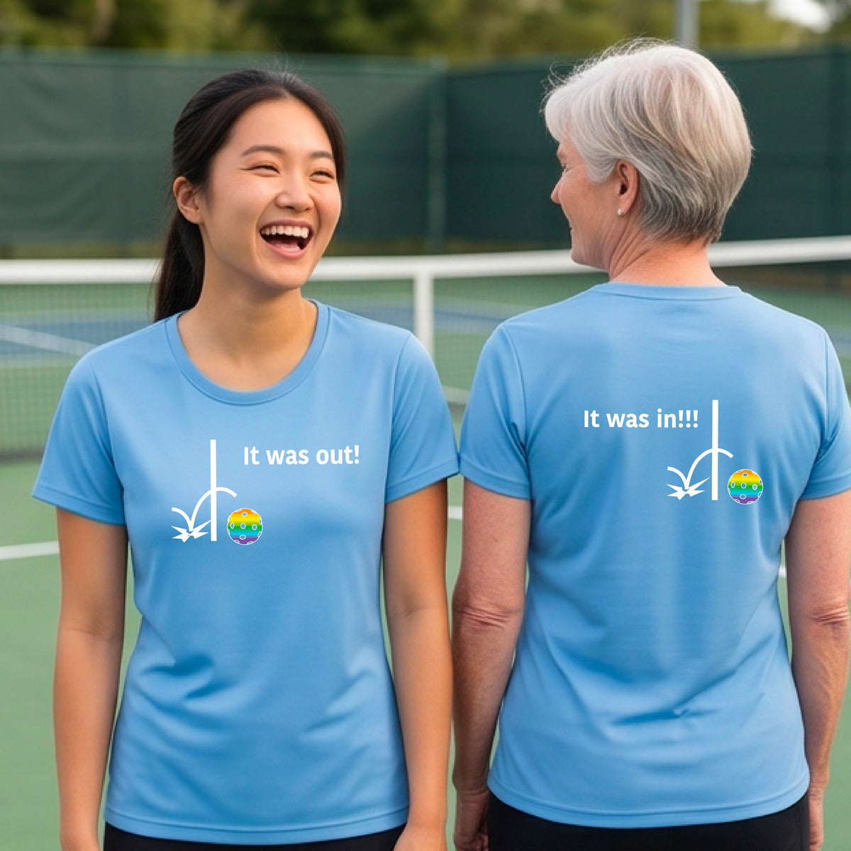 Two women showing a light blue short-sleeve t-shirt with a white design of ‘IT WAS OUT!’ and a colorful pickleball on the front, and the back design with a white ‘IT WAS IN!!!’ and a colorful pickleball, standing on a pickleball court.