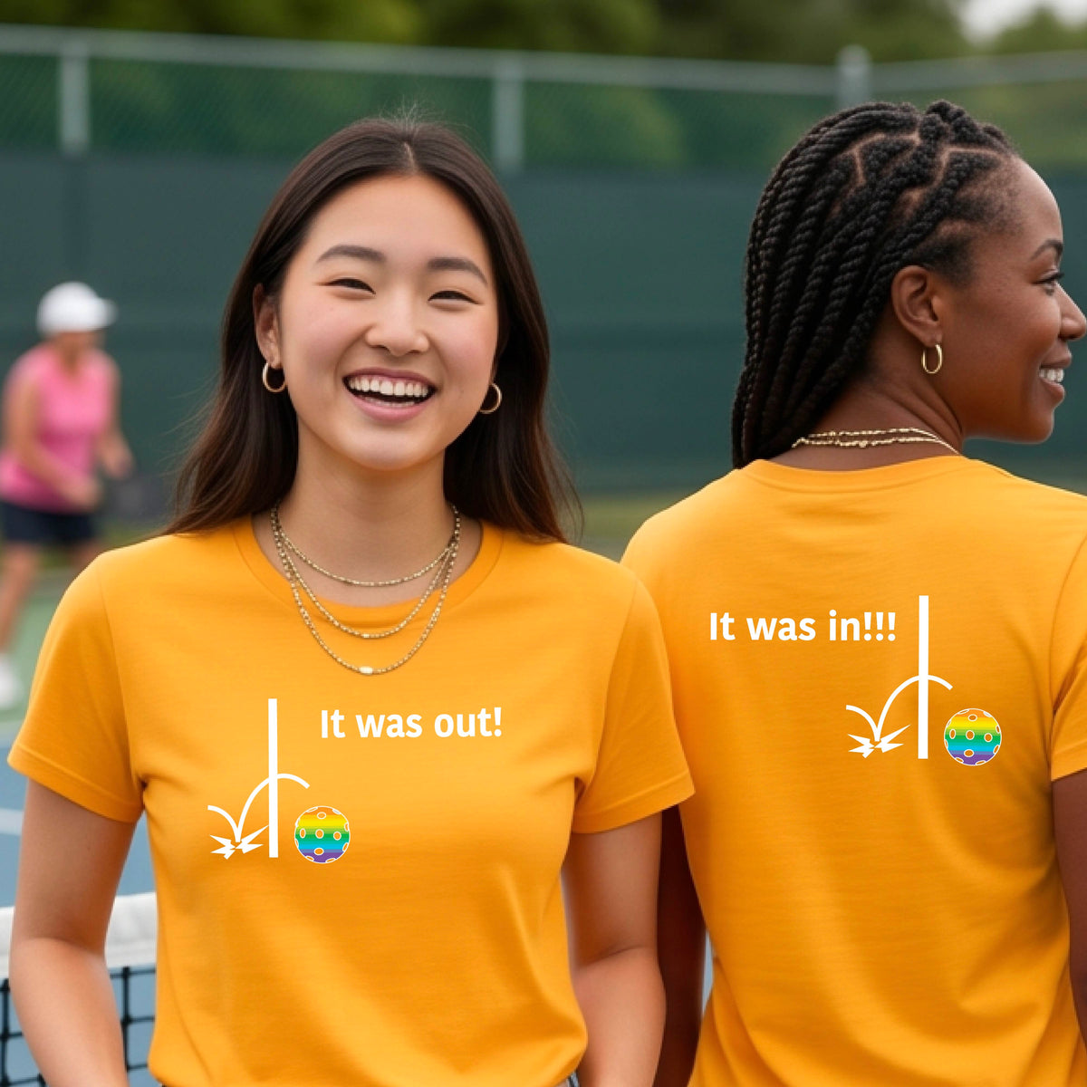Two women showing a mustard yellow gold short-sleeve t-shirt with a white design of ‘IT WAS OUT!’ and a colorful pickleball on the front, and the back design with a white ‘IT WAS IN!!!’ and a colorful pickleball, standing on a pickleball court.