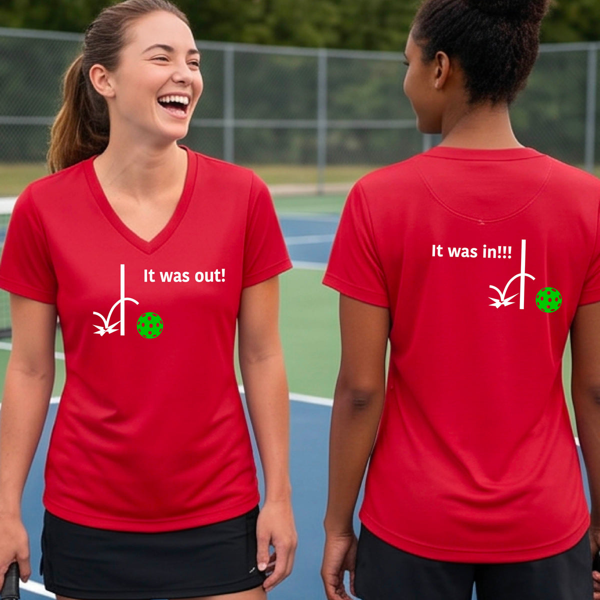 Two women showing a red short-sleeve t-shirt with a white design of ‘IT WAS OUT!’ and a colorful pickleball on the front, and the back design with a white ‘IT WAS IN!!!’ and a colorful pickleball, standing on a pickleball court.