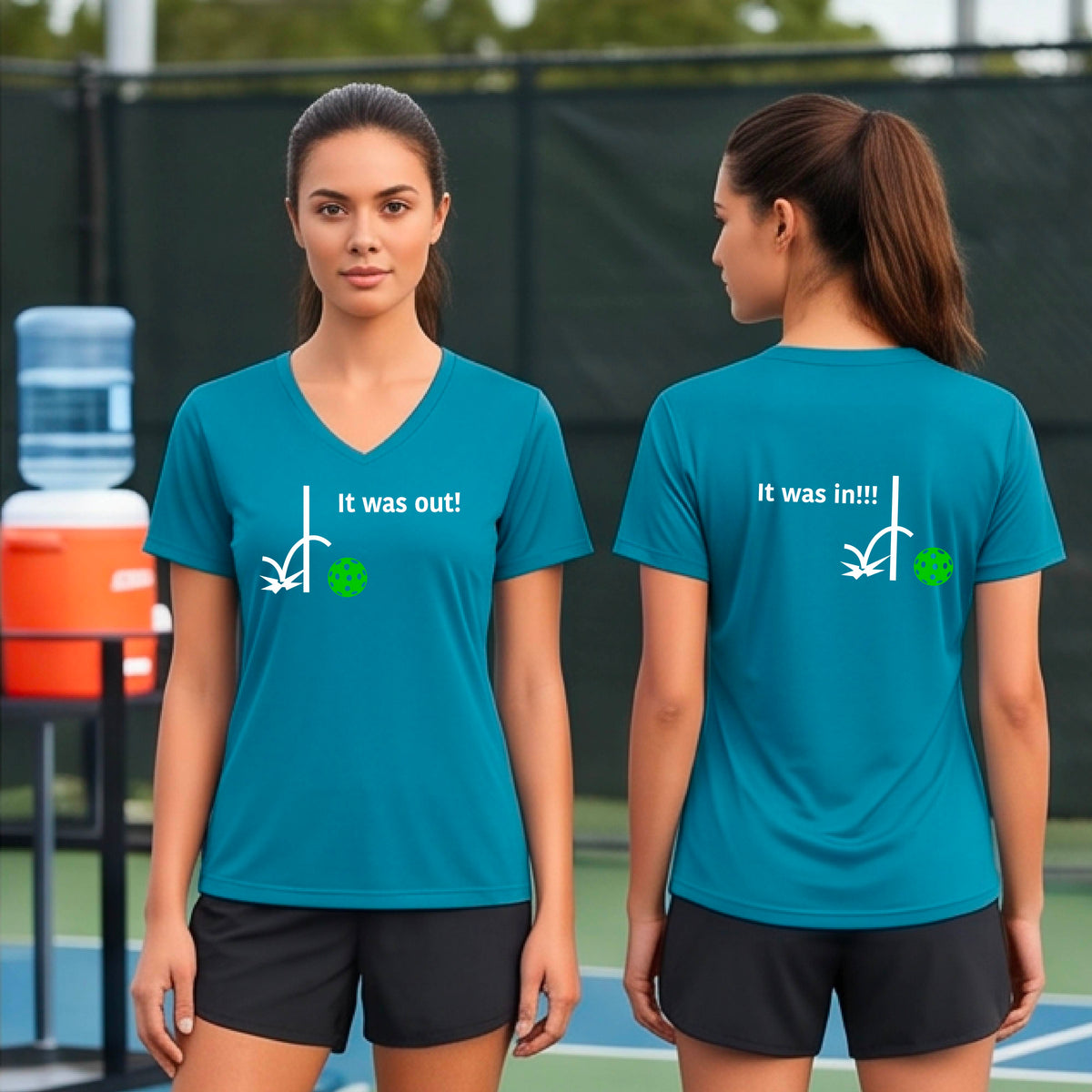 Two women showing a teal short-sleeve t-shirt with a white design of ‘IT WAS OUT!’ and a colorful pickleball on the front, and the back design with a white ‘IT WAS IN!!!’ and a colorful pickleball, standing on a pickleball court.