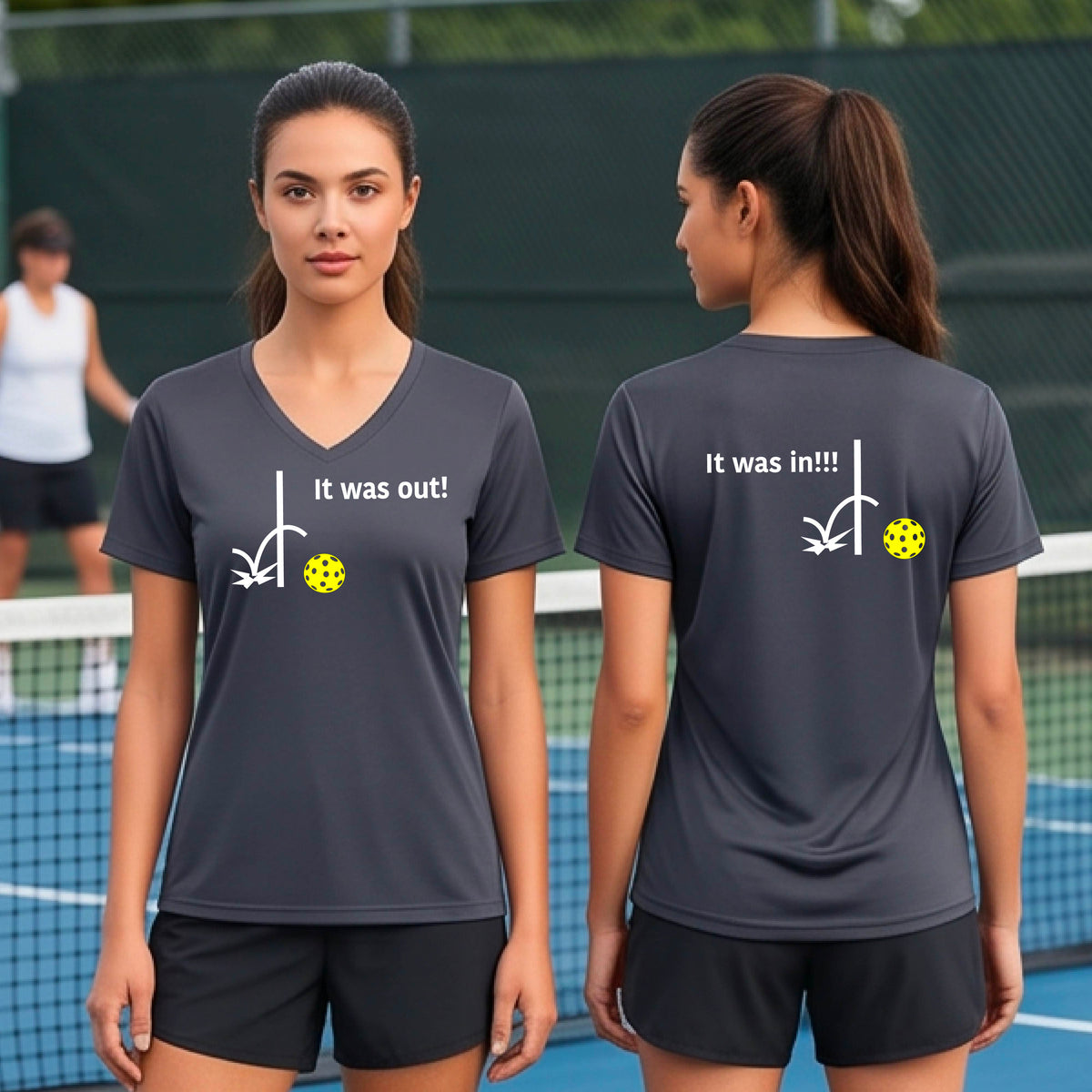 Two women showing a dark gray short-sleeve t-shirt with a white design of ‘IT WAS OUT!’ and a colorful pickleball on the front, and the back design with a white ‘IT WAS IN!!!’ and a colorful pickleball, standing on a pickleball court.