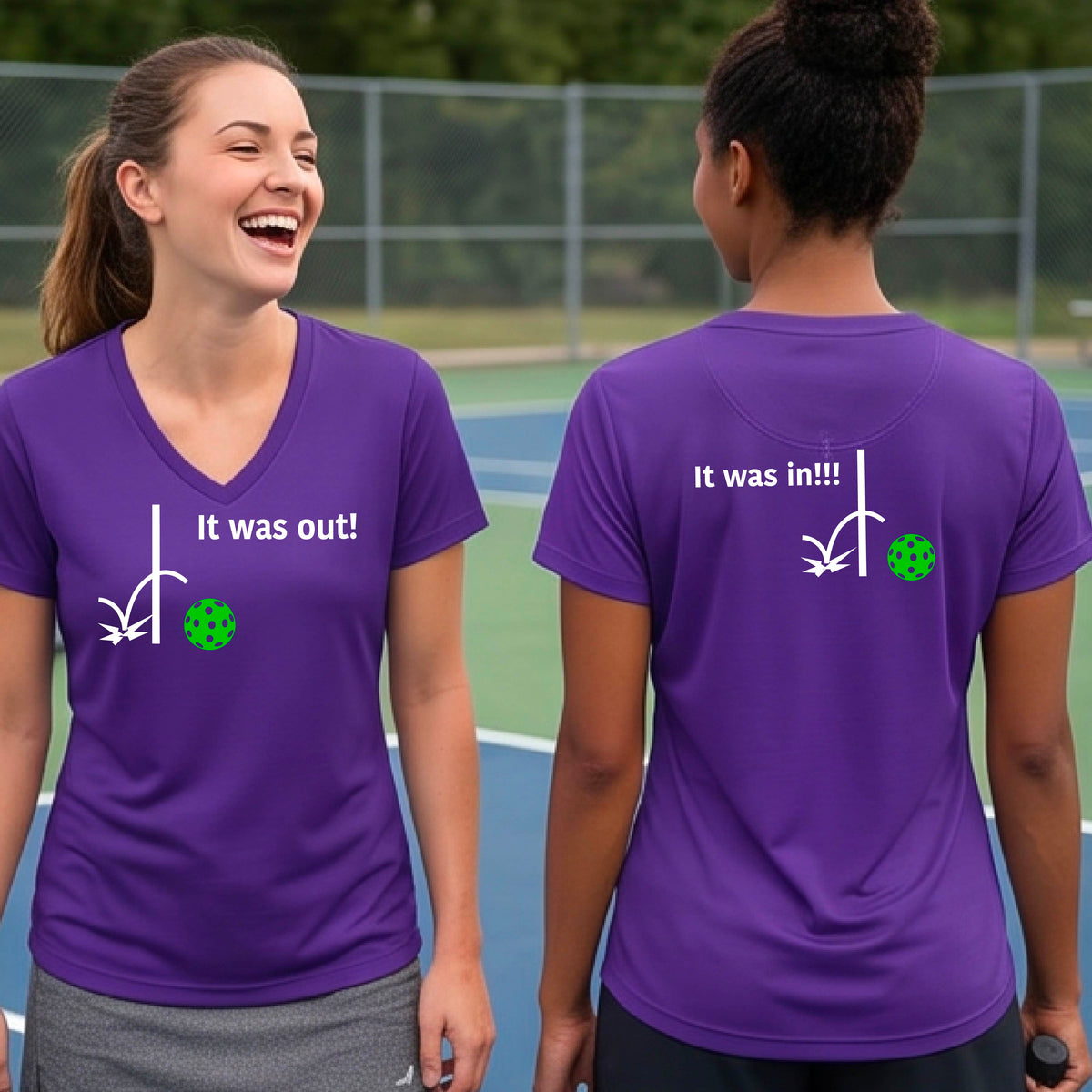 Two women showing a purple short-sleeve t-shirt with a white design of ‘IT WAS OUT!’ and a colorful pickleball on the front, and the back design with a white ‘IT WAS IN!!!’ and a colorful pickleball, standing on a pickleball court.