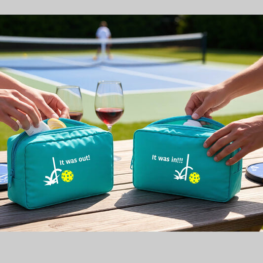 Two turquoise cosmetic toiletry bags with a white design of ‘IT WAS OUT!’ and a colorful pickleball on the front, and the back design with a white ‘IT WAS IN!!!’ and a 