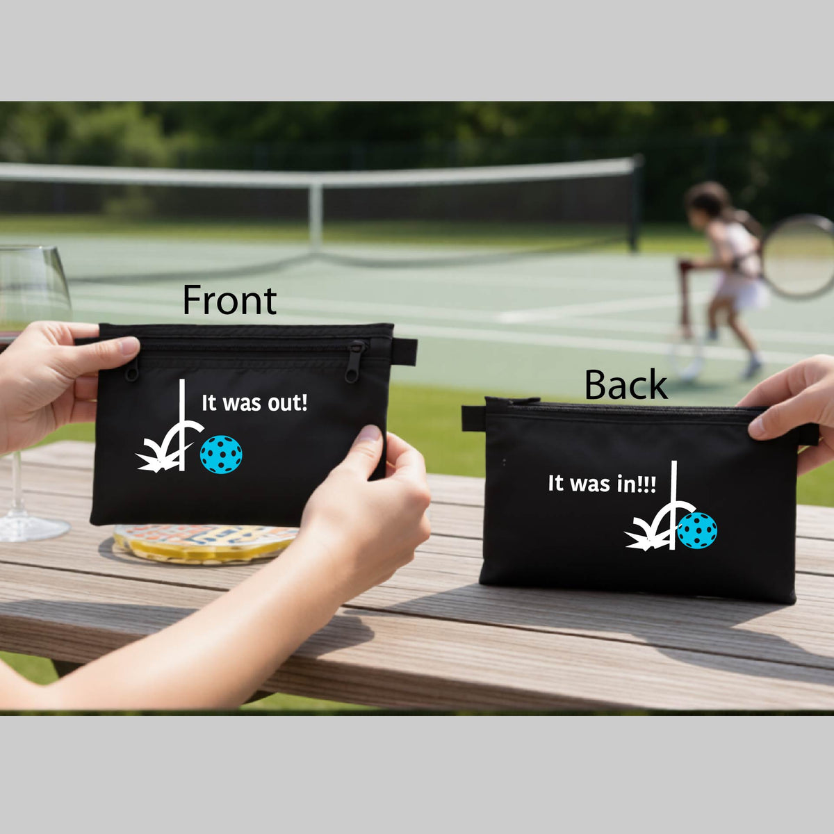A black cosmetic toiletry bag with a white design of ‘IT WAS OUT!’ and a colorful pickleball on the front, and the back design with a white ‘IT WAS IN!!!’ and a colorful pickleball,  with a pickleball court in the background.