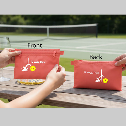 A coral colored cosmetic toiletry bag with a white design of ‘IT WAS OUT!’ and a colorful pickleball on the front, and the back design with a white ‘IT WAS IN!!!’ and a colorful pickleball,  with a pickleball court in the background.