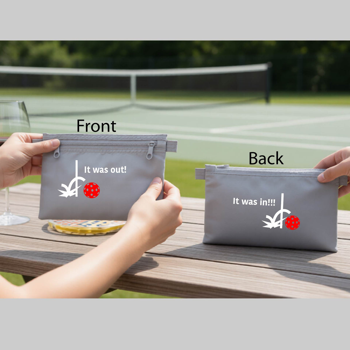 A gray cosmetic toiletry bag with a white design of ‘IT WAS OUT!’ and a colorful pickleball on the front, and the back design with a white ‘IT WAS IN!!!’ and a colorful pickleball,  with a pickleball court in the background.