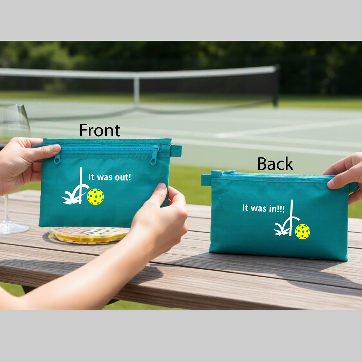A aqua blue cosmetic toiletry bag with a white design of ‘IT WAS OUT!’ and a colorful pickleball on the front, and the back design with a white ‘IT WAS IN!!!’ and a colorful pickleball,  with a pickleball court in the background.