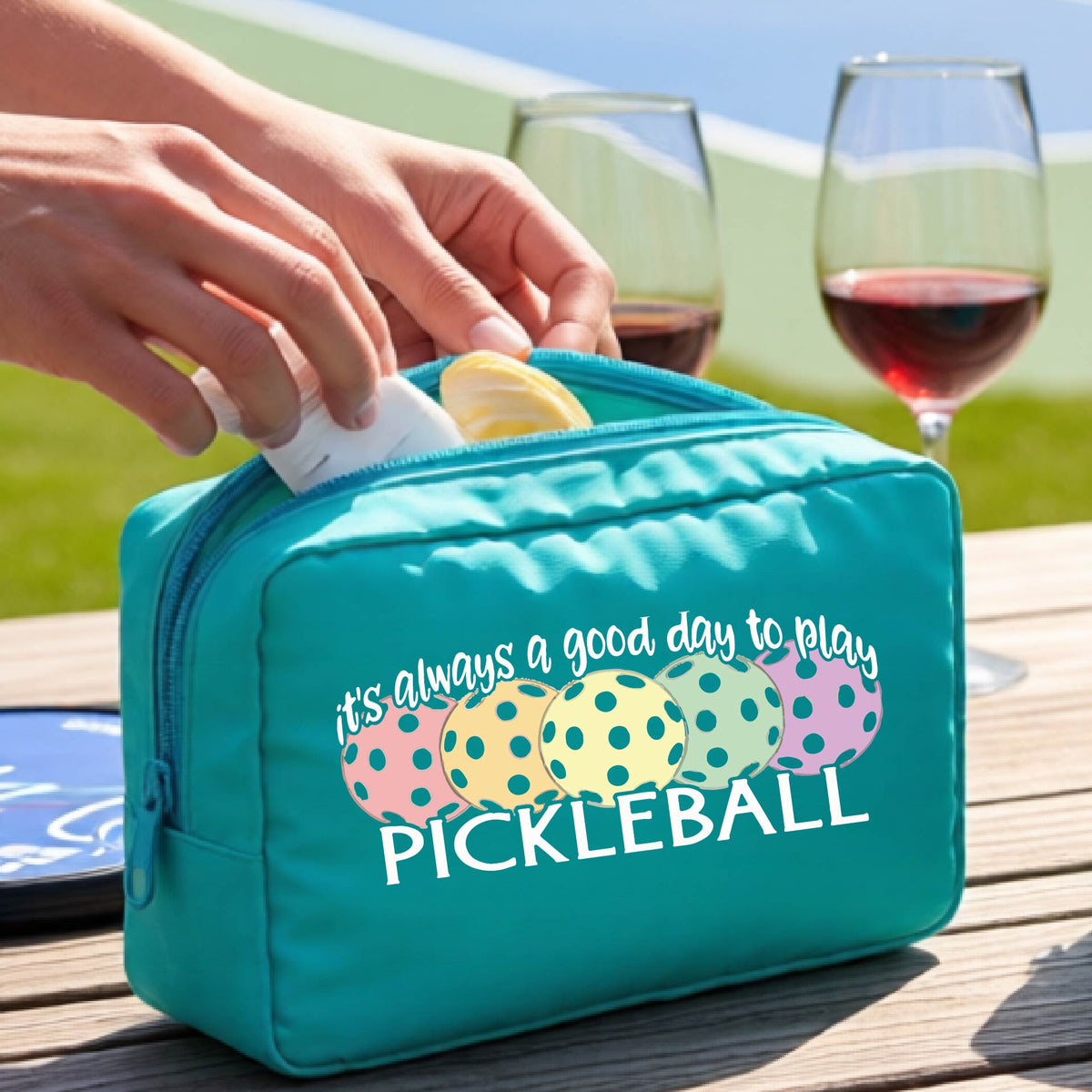 A turquoise pickleball-themed zippered cosmetic pouch with a printed graphic and the phrase 'it's always a good day to play PICKLEBALL' .
