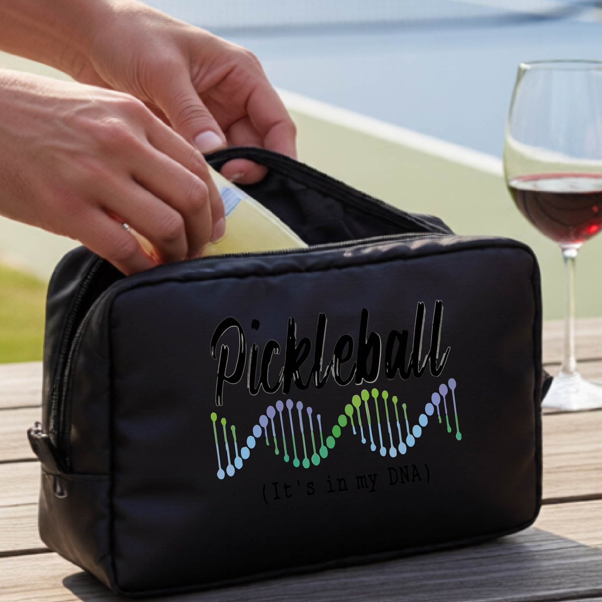 A black zippered cosmetic pouch bag with 'Pickleball (It's in my DNA)' printed on the front, at an outdoor table and a pickleball court in the background,. 