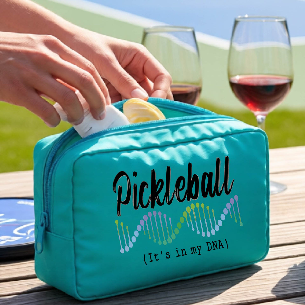 A turquoise zippered cosmetic pouch bag with 'Pickleball (It's in my DNA)' printed on the front, at an outdoor table and a pickleball court in the background,. 