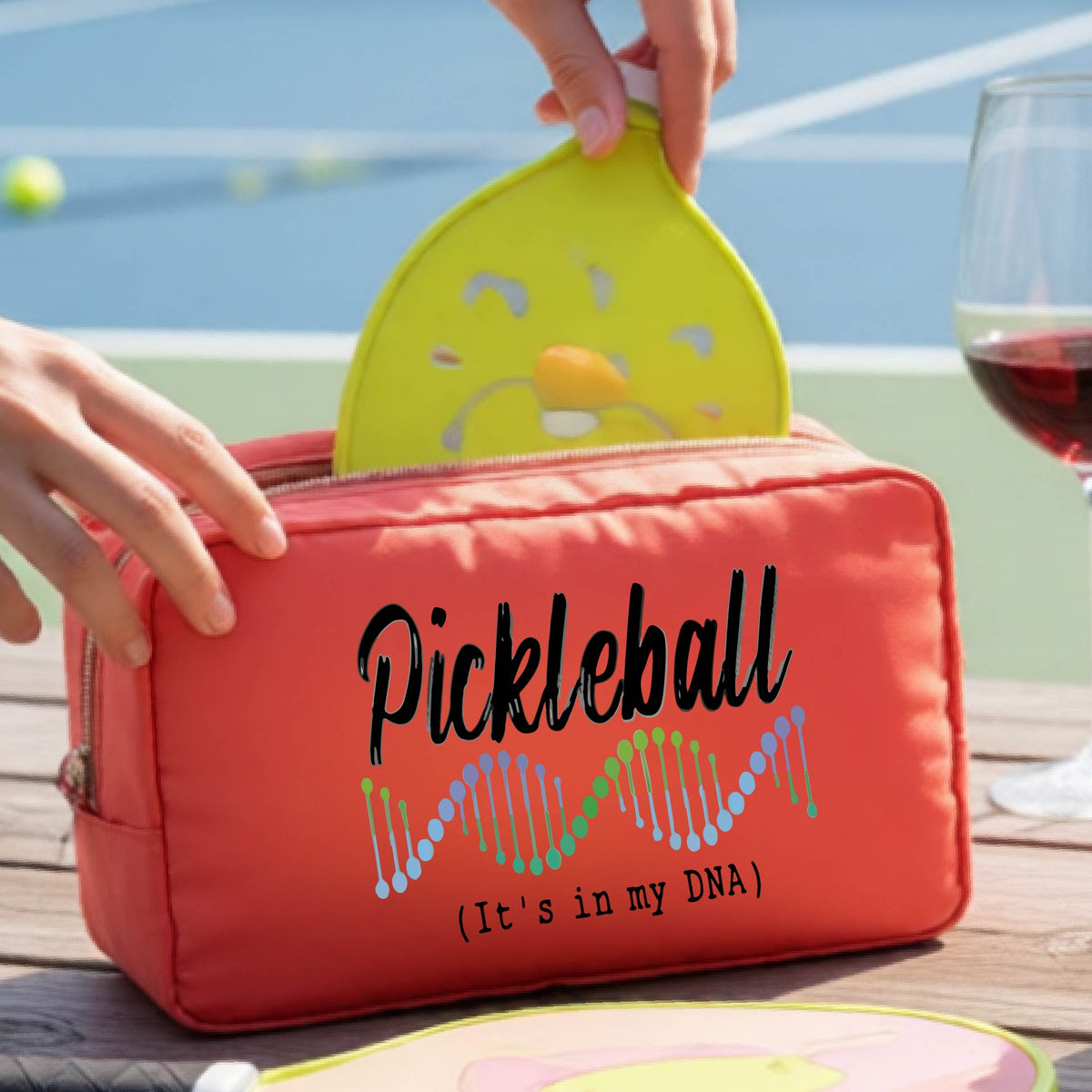 A coral colored zippered cosmetic pouch bag with 'Pickleball (It's in my DNA)' printed on the front, at an outdoor table and a pickleball court in the background,. 