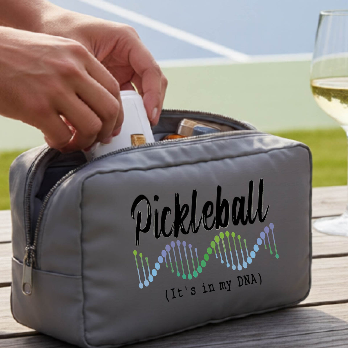 A gray zippered cosmetic pouch bag with 'Pickleball (It's in my DNA)' printed on the front, at an outdoor table and a pickleball court in the background,. 