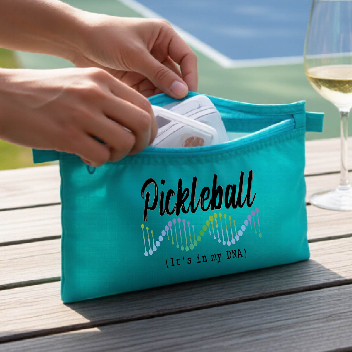 A turquoise blue pickleball-themed zippered cosmetic pouch on a wooden table, with a pickleball court in the background. 