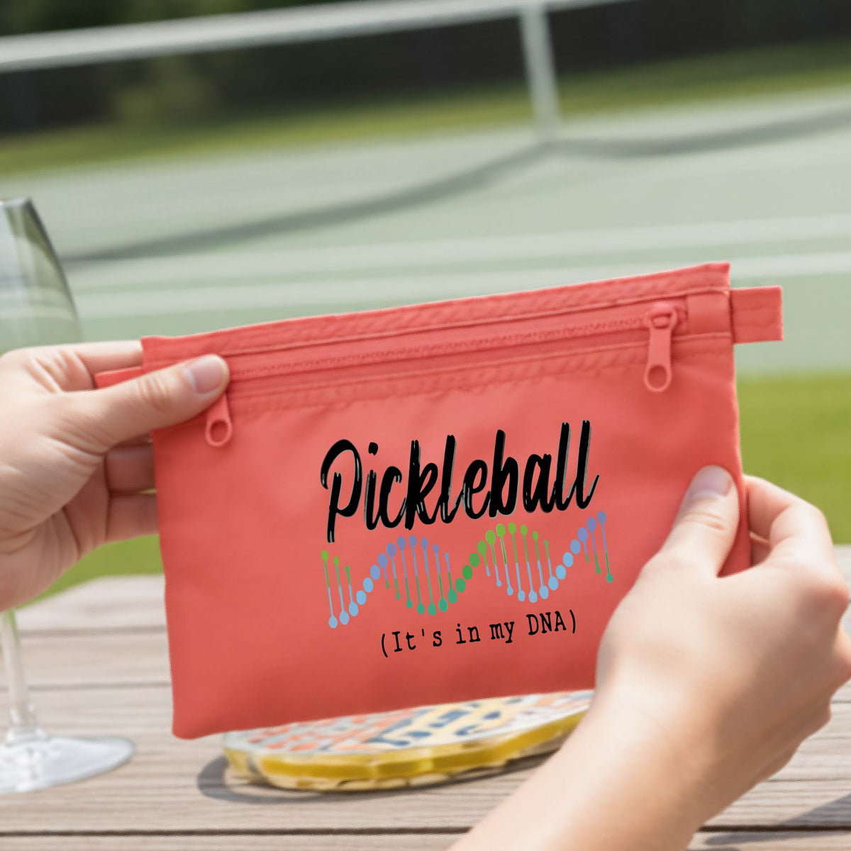 A coral-colored blue pickleball-themed zippered cosmetic pouch on a wooden table, with a pickleball court in the background. 