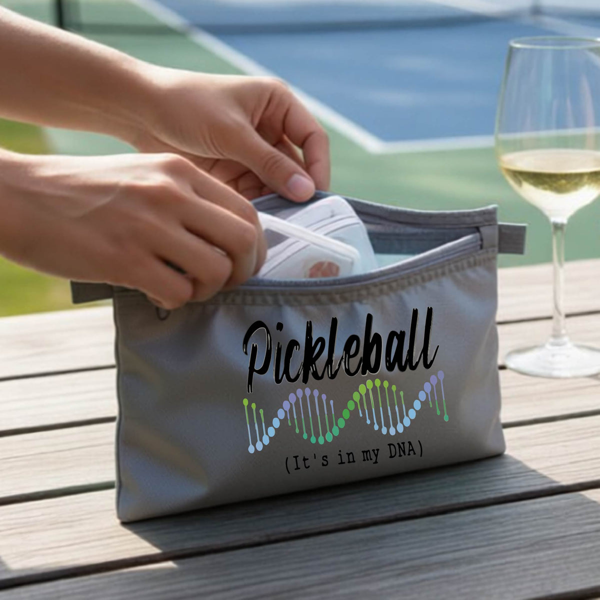 A gray pickleball-themed zippered cosmetic pouch on a wooden table, with a pickleball court in the background. 