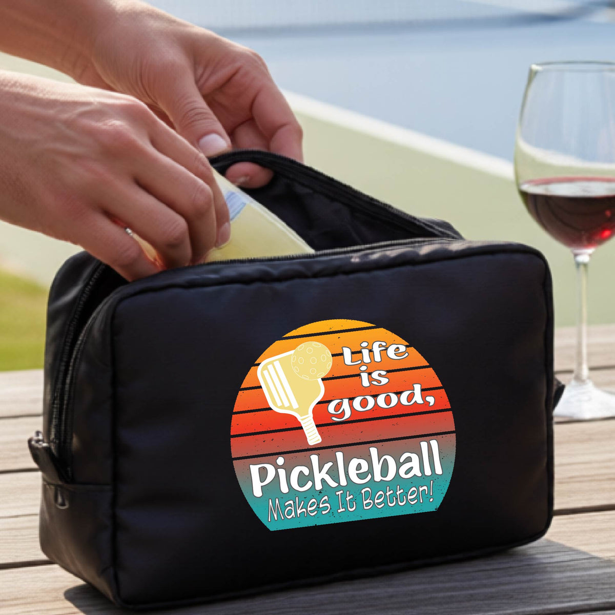 A black zippered pickleball-themed toiletry bag sits on a wooden table outdoors, with a person's hands placing a bottle inside. The bag features a colorful retro-style circular graphic with a pickleball paddle and the text 'Life is good, Pickleball Makes It Better!'.
