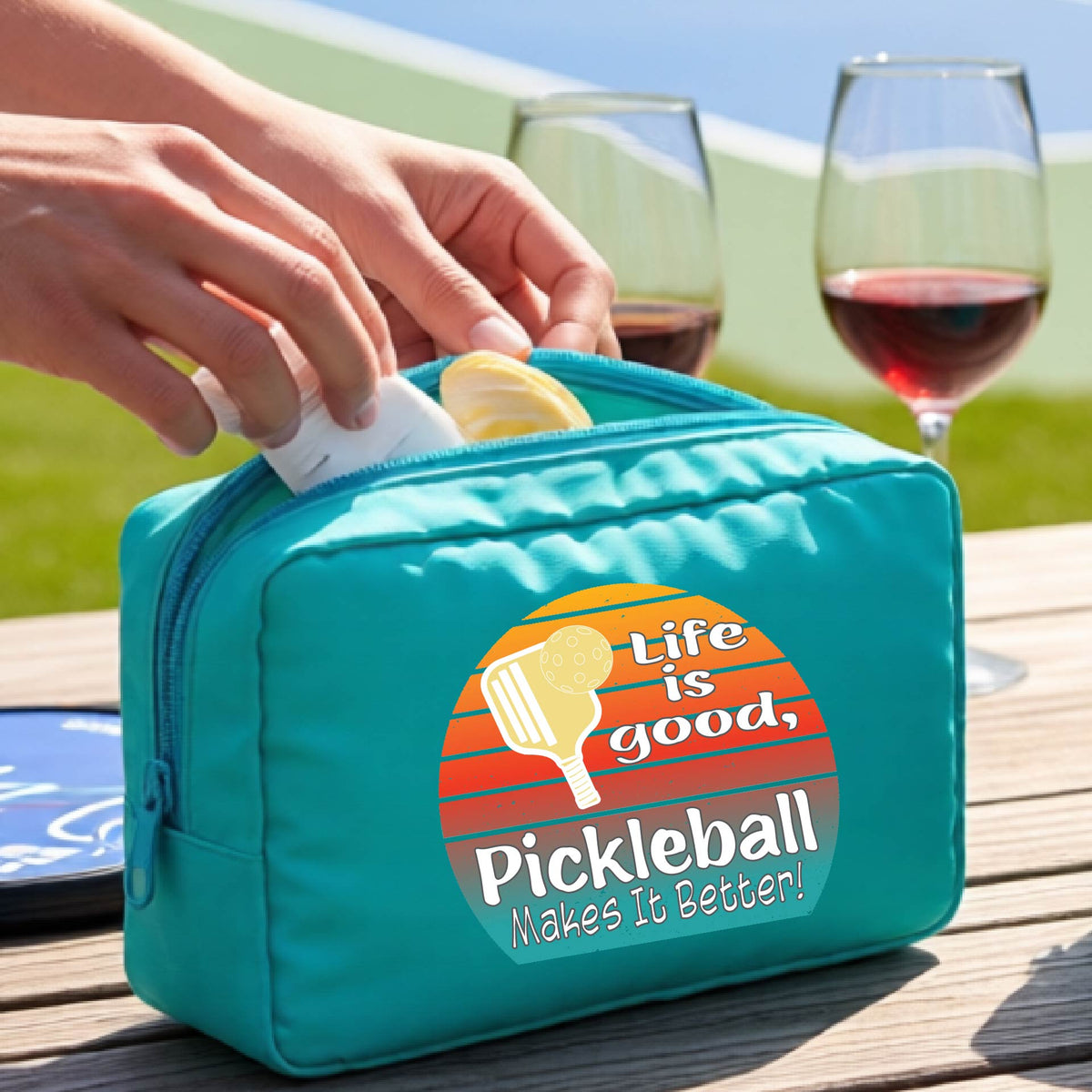 A turquoise blue zippered pickleball-themed toiletry bag sits on a wooden table outdoors, with a person's hands placing a bottle inside. The bag features a colorful retro-style circular graphic with a pickleball paddle and the text 'Life is good, Pickleball Makes It Better!'.