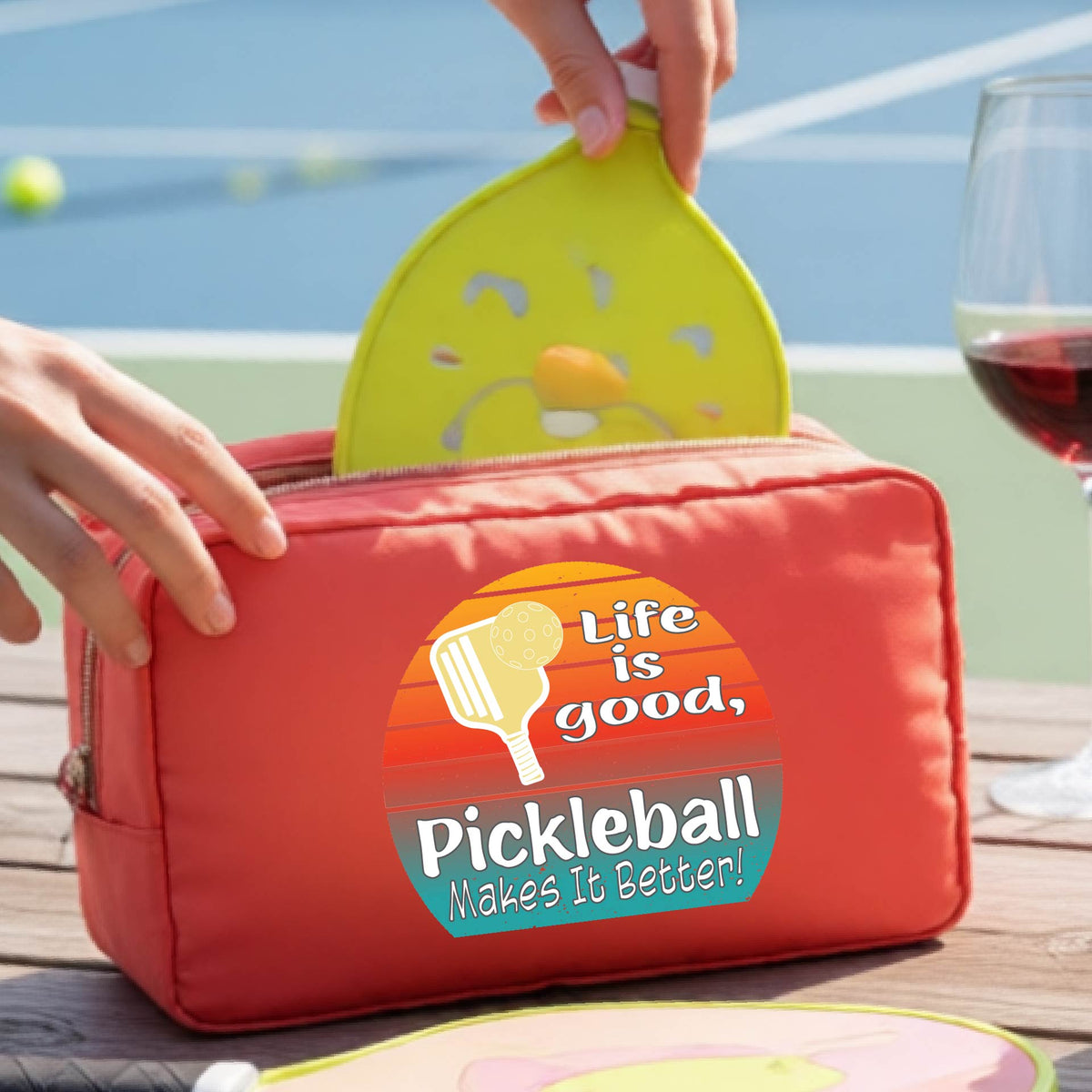 A coral orange zippered pickleball-themed toiletry bag sits on a wooden table outdoors, with a person's hands placing a bottle inside. The bag features a colorful retro-style circular graphic with a pickleball paddle and the text 'Life is good, Pickleball Makes It Better!'.
