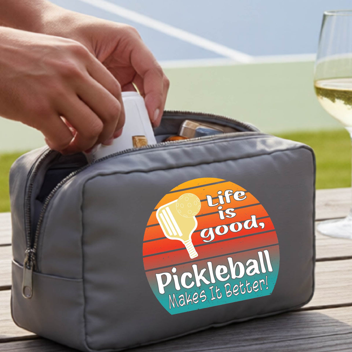 A gray zippered pickleball-themed toiletry bag sits on a wooden table outdoors, with a person's hands placing a bottle inside. The bag features a colorful retro-style circular graphic with a pickleball paddle and the text 'Life is good, Pickleball Makes It Better!'.