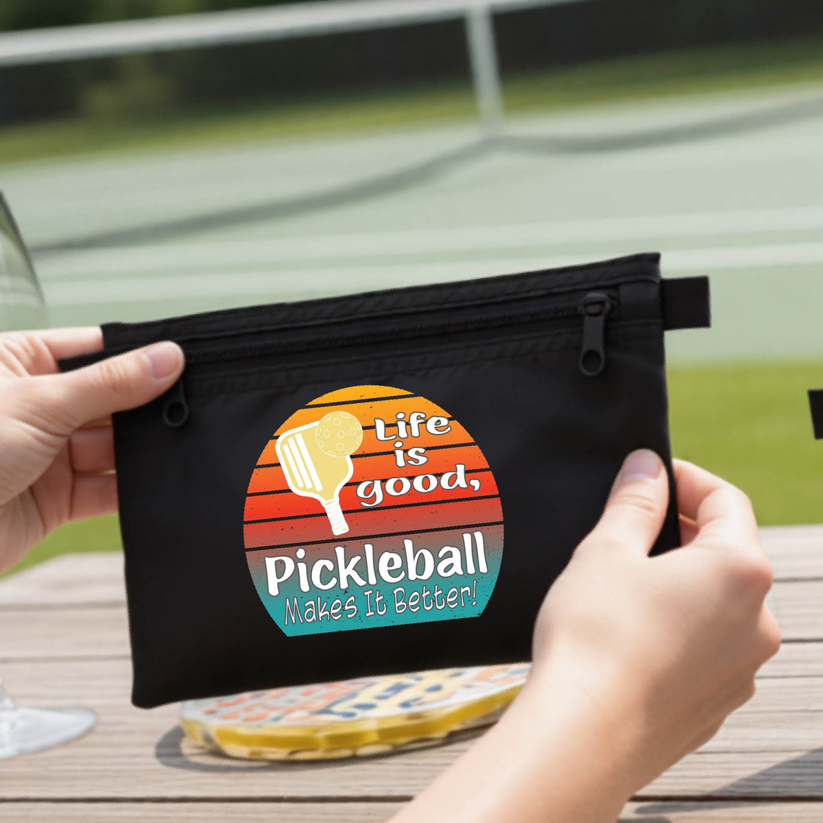 A black zippered pouch held by two hands, featuring a circular retro pickleball graphic with the text 'Life is good, Pickleball Makes It Better!'. The background shows a blurred outdoor pickleball court and a wooden table.
