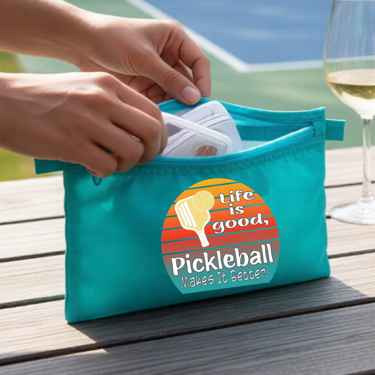 A turquoise blue zippered pouch held by two hands, featuring a circular retro pickleball graphic with the text 'Life is good, Pickleball Makes It Better!'. The background shows a blurred outdoor pickleball court and a wooden table.