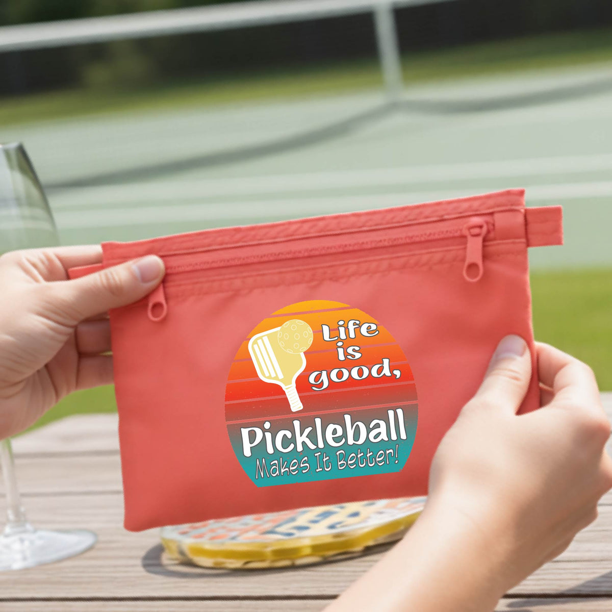 A coral orange zippered pouch held by two hands, featuring a circular retro pickleball graphic with the text 'Life is good, Pickleball Makes It Better!'. The background shows a blurred outdoor pickleball court and a wooden table.