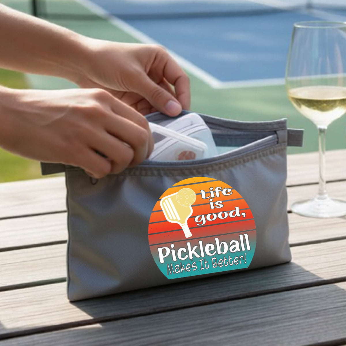 A gray zippered pouch held by two hands, featuring a circular retro pickleball graphic with the text 'Life is good, Pickleball Makes It Better!'. The background shows a blurred outdoor pickleball court and a wooden table.