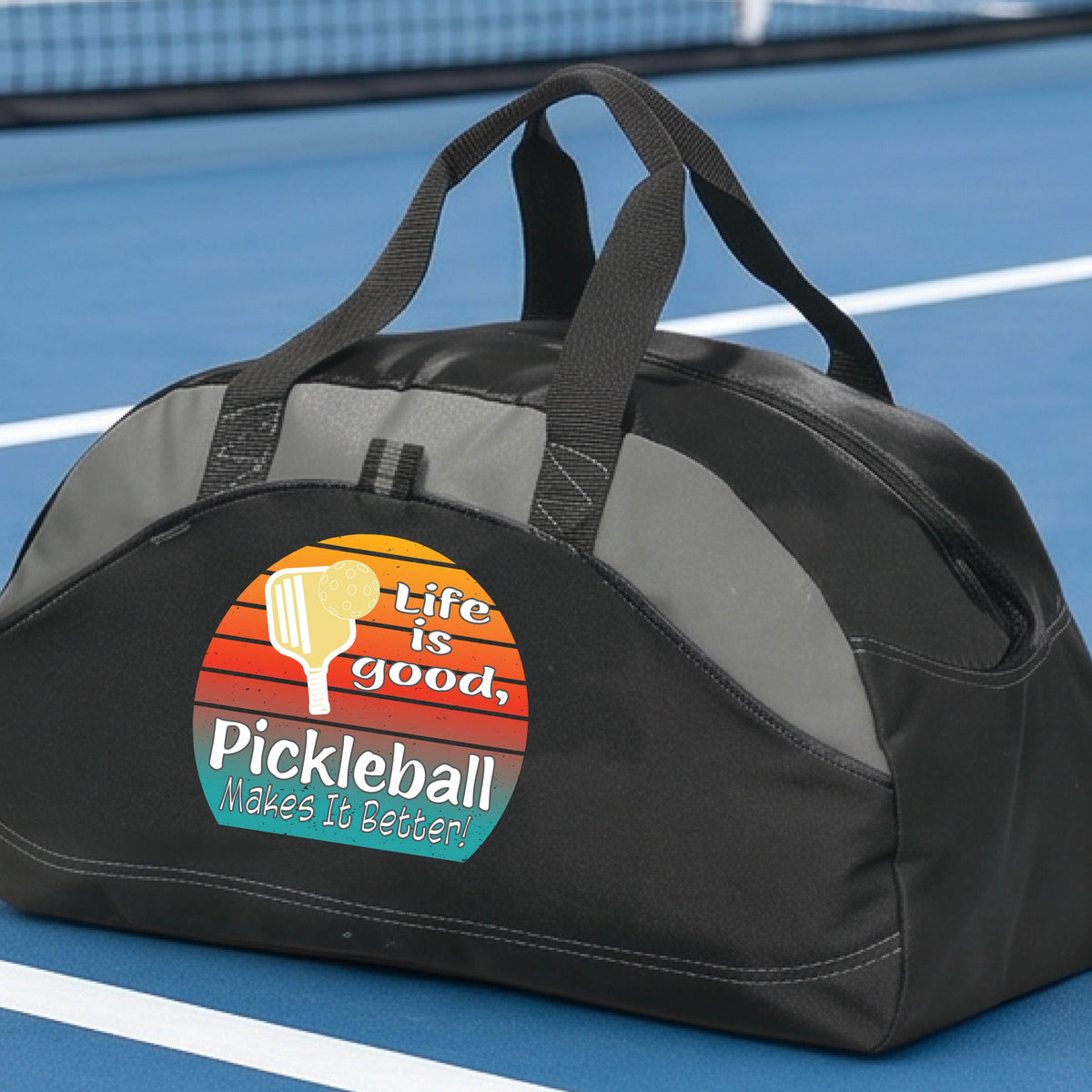 A gray and black mediium size duffel bag with a retro multi-colored circle with a pickleball paddle and ball, with white text that says 'LIFE IS GOOD PICKLEBALL MAKES IT BETTER', standing in front of a pickleball court.