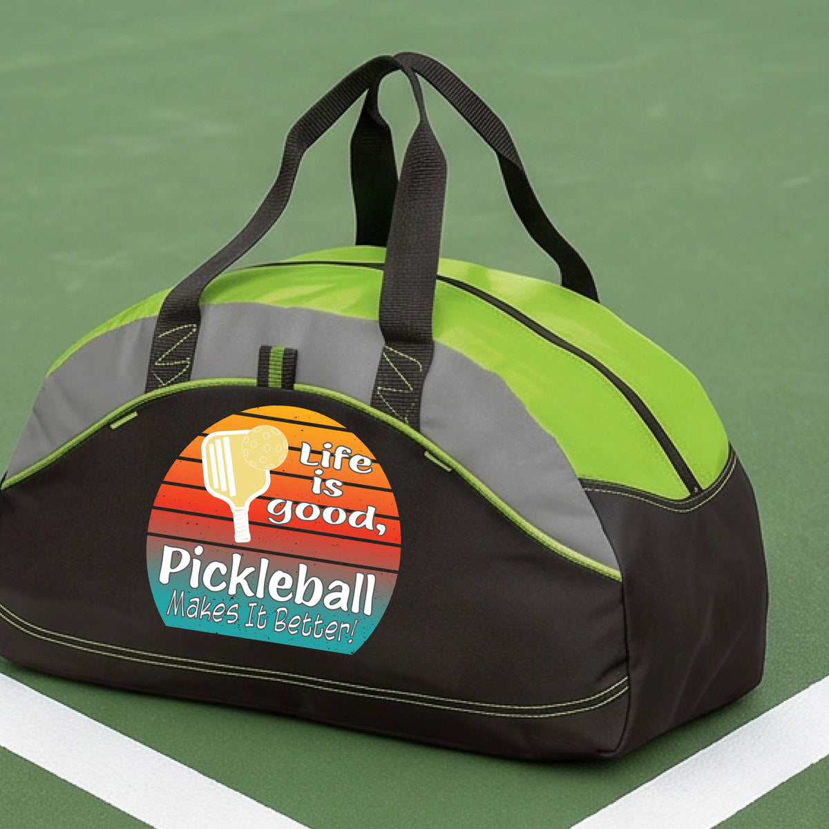 A lime green, gray and black mediium size duffel bag with a retro multi-colored circle with a pickleball paddle and ball, with white text that says 'LIFE IS GOOD PICKLEBALL MAKES IT BETTER', standing in front of a pickleball court.