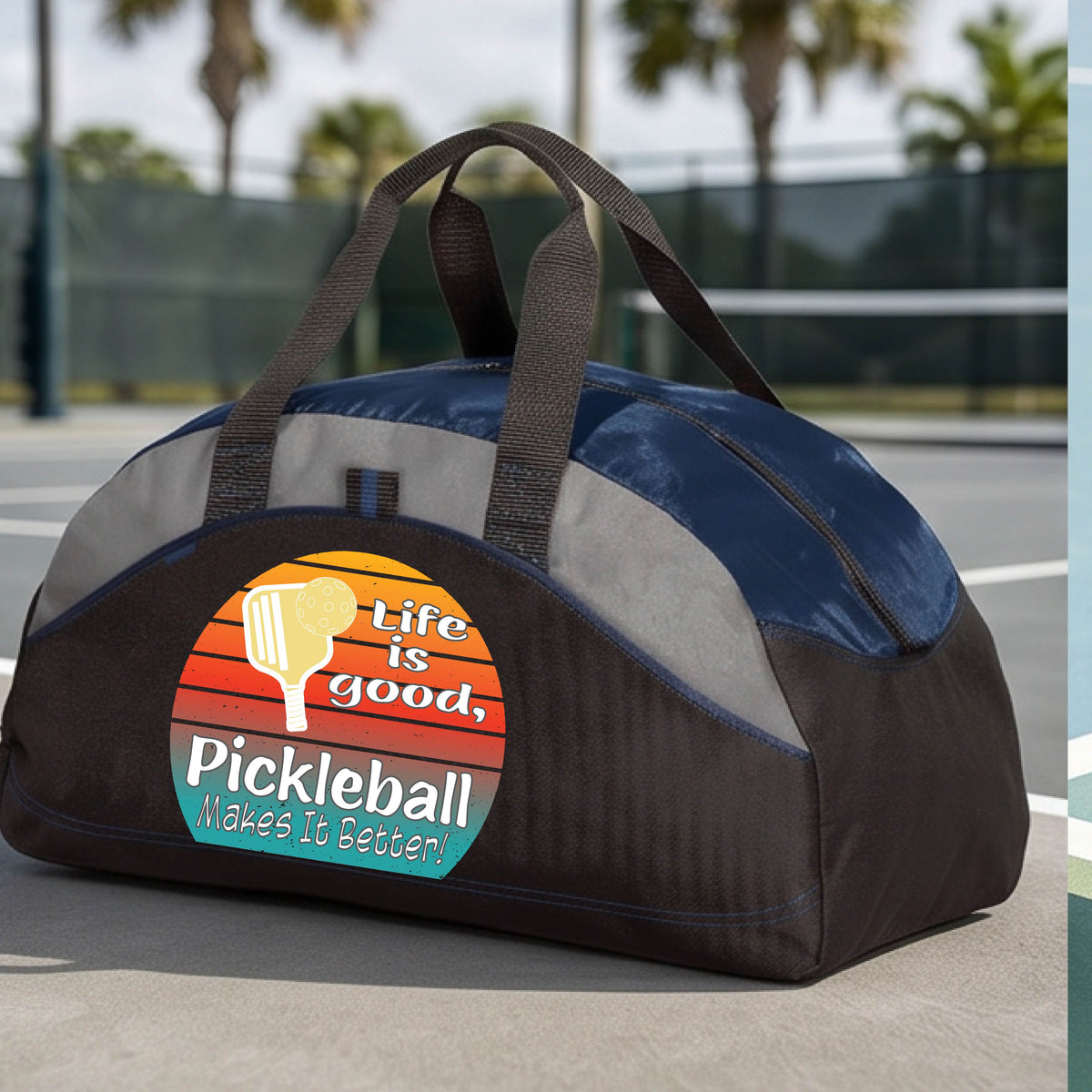 A navy blue, gray and black mediium size duffel bag with a retro multi-colored circle with a pickleball paddle and ball, with white text that says 'LIFE IS GOOD PICKLEBALL MAKES IT BETTER', standing in front of a pickleball court.