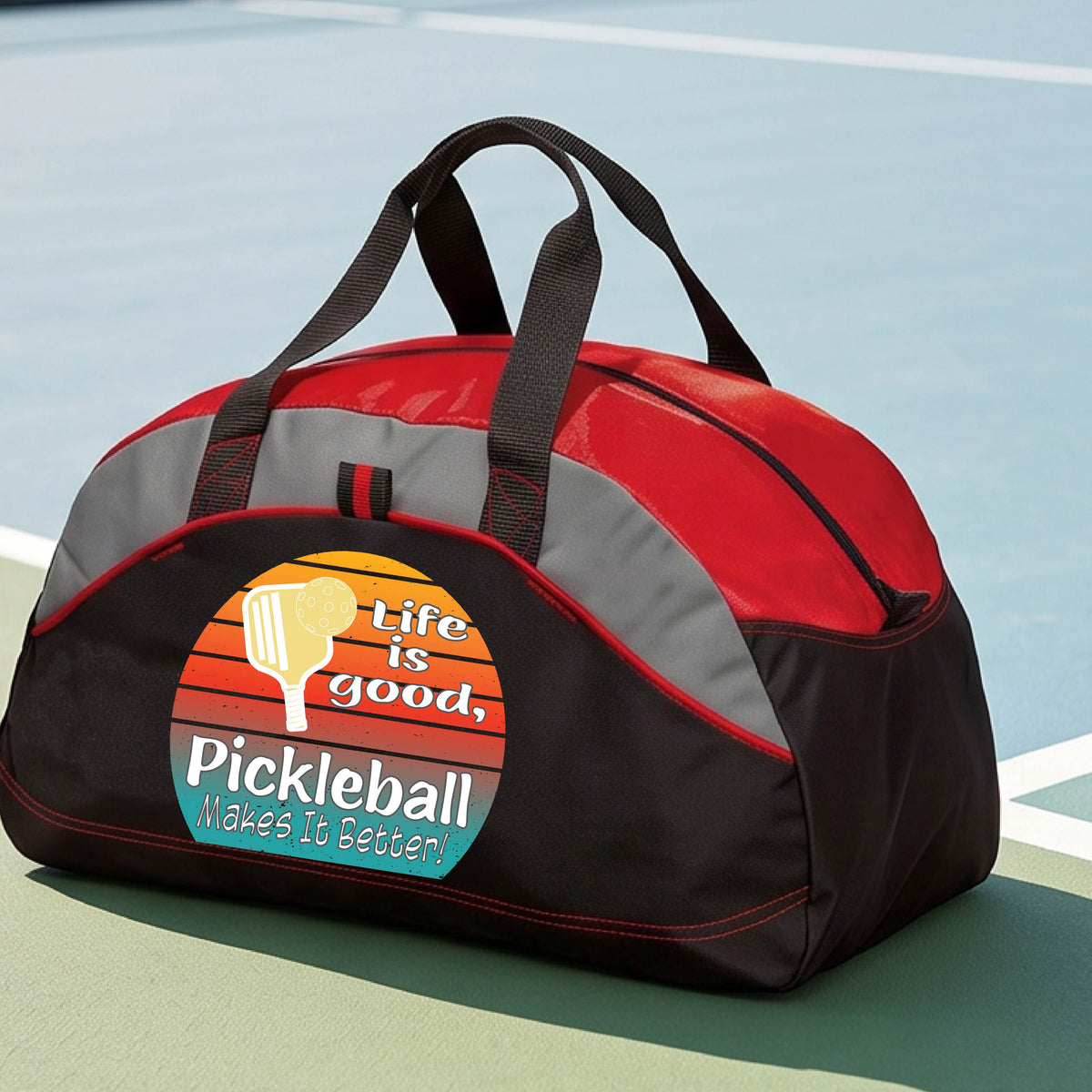 A red, gray and black mediium size duffel bag with a retro multi-colored circle with a pickleball paddle and ball, with white text that says 'LIFE IS GOOD PICKLEBALL MAKES IT BETTER', standing in front of a pickleball court.