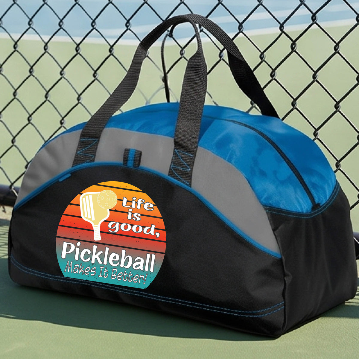 A royal blue, gray and black mediium size duffel bag with a retro multi-colored circle with a pickleball paddle and ball, with white text that says 'LIFE IS GOOD PICKLEBALL MAKES IT BETTER', standing in front of a pickleball court.