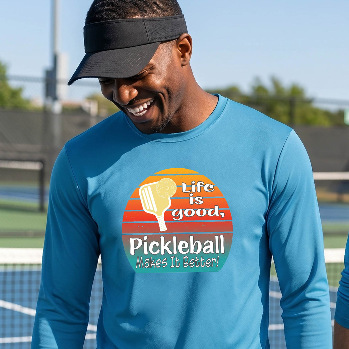 A man wearing a bright blue long-sleeve t-shirt with a retro multi-colored circle with a pickleball paddle and ball, with white text that says 'LIFE IS GOOD PICKLEBALL MAKES IT BETTER', standing in front of a pickleball court.
