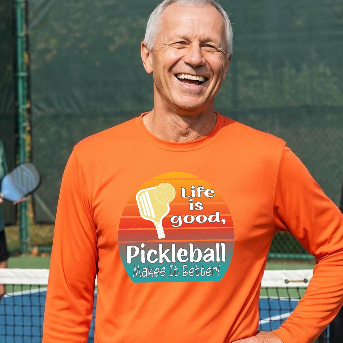 A man wearing an orange long-sleeve t-shirt with a retro multi-colored circle with a pickleball paddle and ball, with white text that says 'LIFE IS GOOD PICKLEBALL MAKES IT BETTER', standing in front of a pickleball court.
