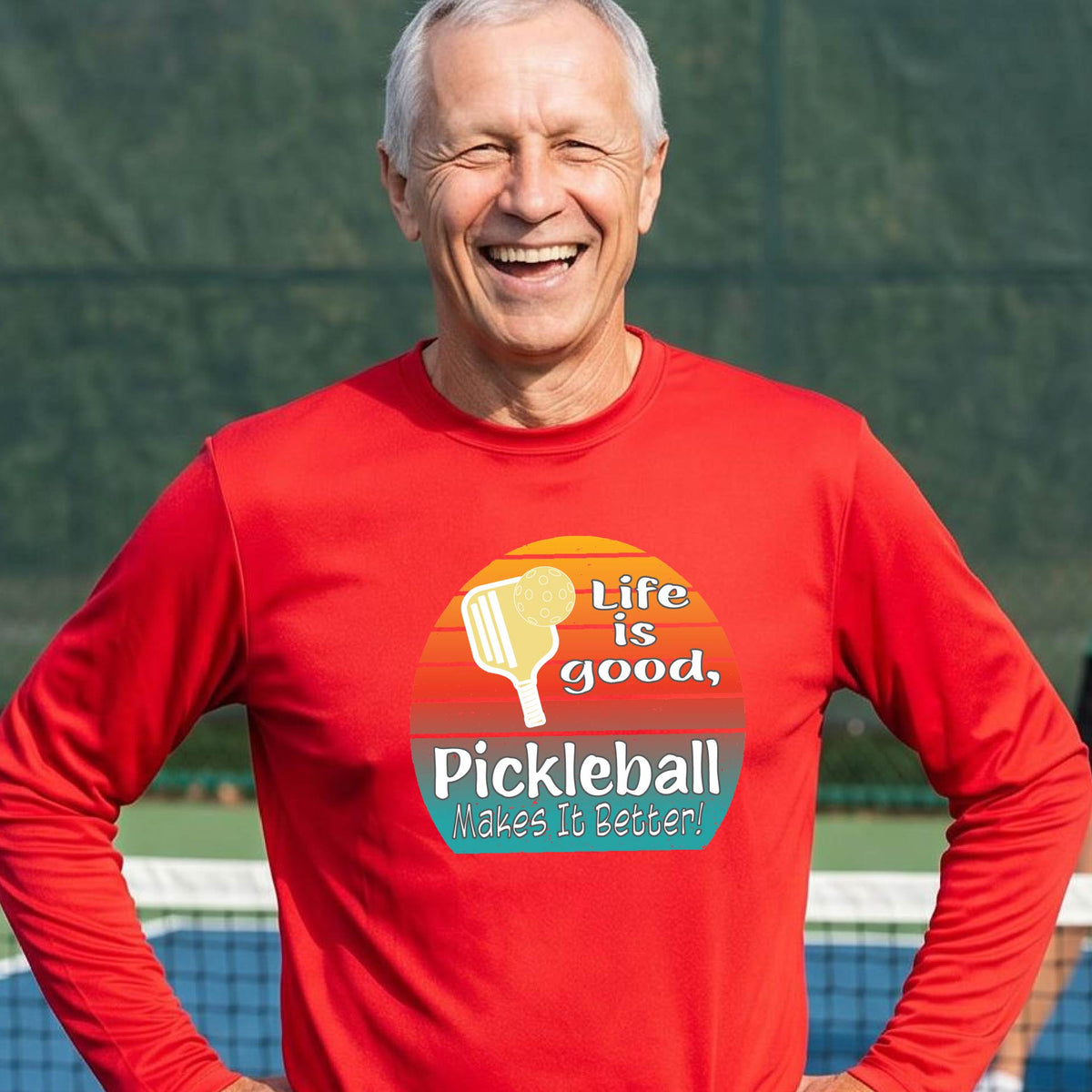 A man wearing a red long-sleeve t-shirt with a retro multi-colored circle with a pickleball paddle and ball, with white text that says 'LIFE IS GOOD PICKLEBALL MAKES IT BETTER', standing in front of a pickleball court.