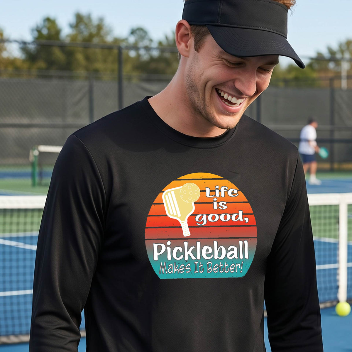 A man wearing a black long-sleeve t-shirt with a retro multi-colored circle with a pickleball paddle and ball, with white text that says 'LIFE IS GOOD PICKLEBALL MAKES IT BETTER', standing in front of a pickleball court.