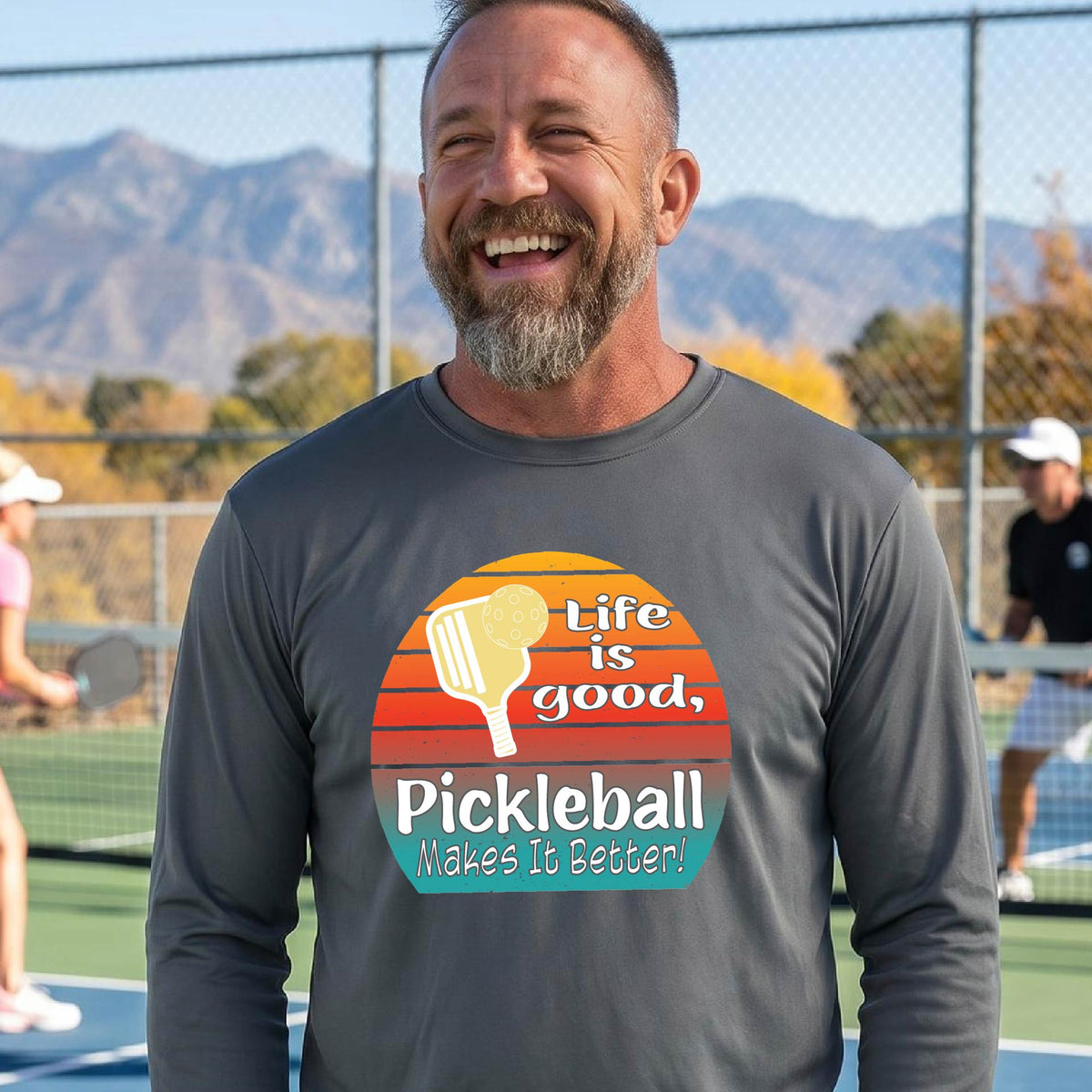 A man wearing a dark gray long-sleeve t-shirt with a retro multi-colored circle with a pickleball paddle and ball, with white text that says 'LIFE IS GOOD PICKLEBALL MAKES IT BETTER', standing in front of a pickleball court.