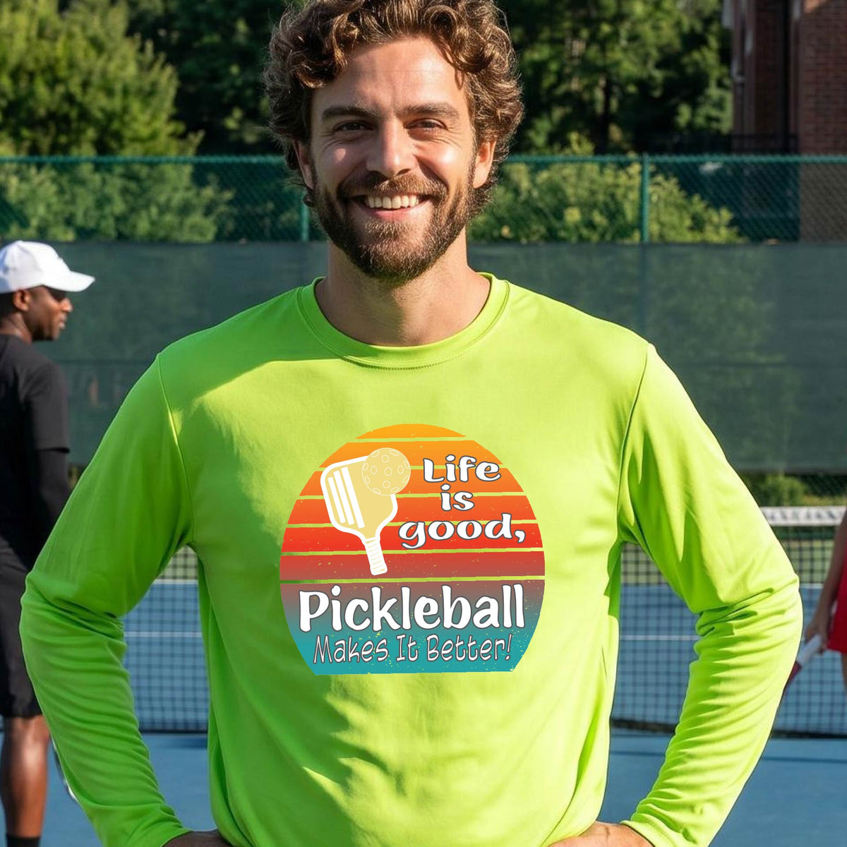 A man wearing a lime green long-sleeve t-shirt with a retro multi-colored circle with a pickleball paddle and ball, with white text that says 'LIFE IS GOOD PICKLEBALL MAKES IT BETTER', standing in front of a pickleball court.