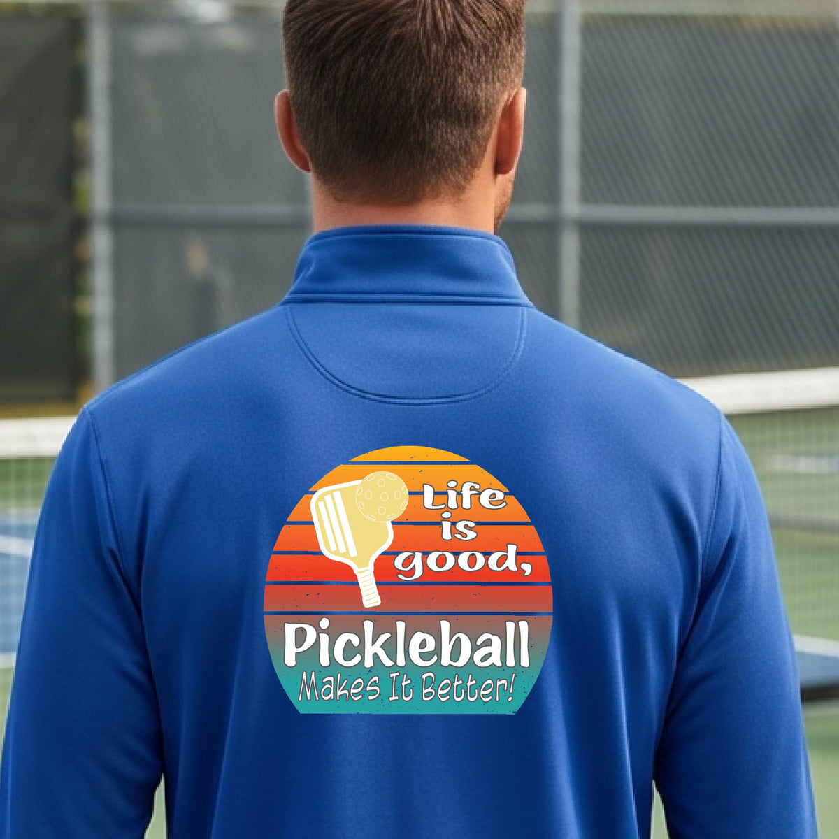A man viewed from behind on an outdoor pickleball court, wearing a royal blue long-sleeve quarter-zip shirt with a large colorful graphic on the back. The graphic features a retro sunset design with a pickleball paddle and the text 'Life is good, Pickleball Makes It Better!'.