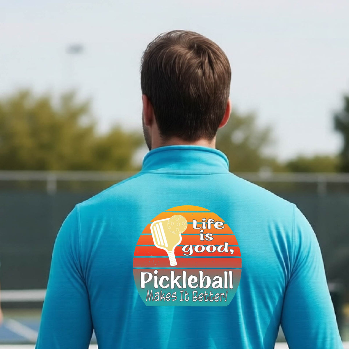 A man viewed from behind on an outdoor pickleball court, wearing a bright blue long-sleeve quarter-zip shirt with a large colorful graphic on the back. The graphic features a retro sunset design with a pickleball paddle and the text 'Life is good, Pickleball Makes It Better!'.