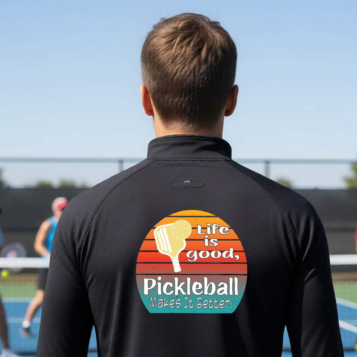 A man viewed from behind on an outdoor pickleball court, wearing a black long-sleeve quarter-zip shirt with a large colorful graphic on the back. The graphic features a retro sunset design with a pickleball paddle and the text 'Life is good, Pickleball Makes It Better!'.