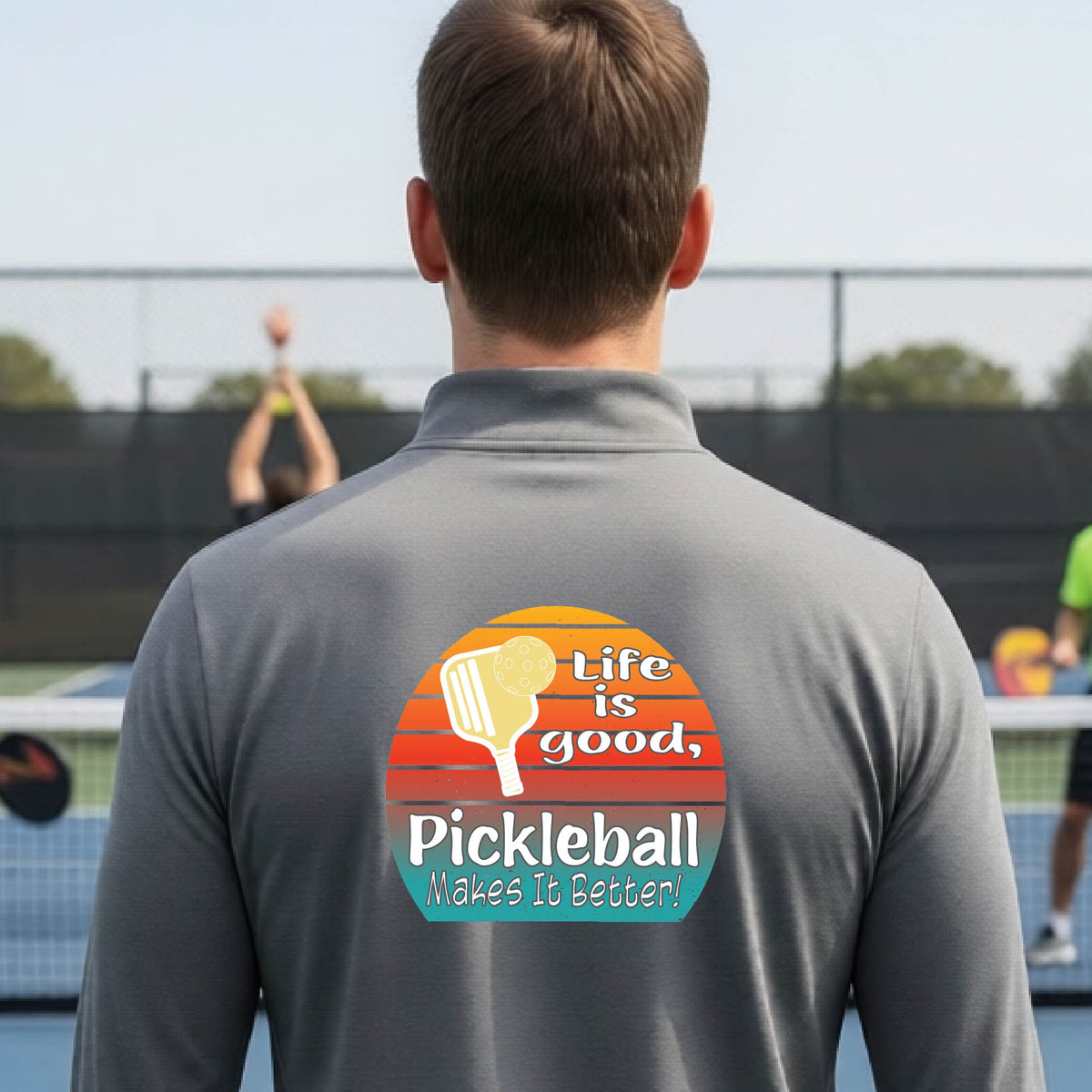 A man viewed from behind on an outdoor pickleball court, wearing a gray long-sleeve quarter-zip shirt with a large colorful graphic on the back. The graphic features a retro sunset design with a pickleball paddle and the text 'Life is good, Pickleball Makes It Better!'.