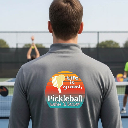 A man viewed from behind on an outdoor pickleball court, wearing a gray long-sleeve quarter-zip shirt with a large colorful graphic on the back. The graphic features a retro sunset design with a pickleball paddle and the text 'Life is good, Pickleball Makes It Better!'.