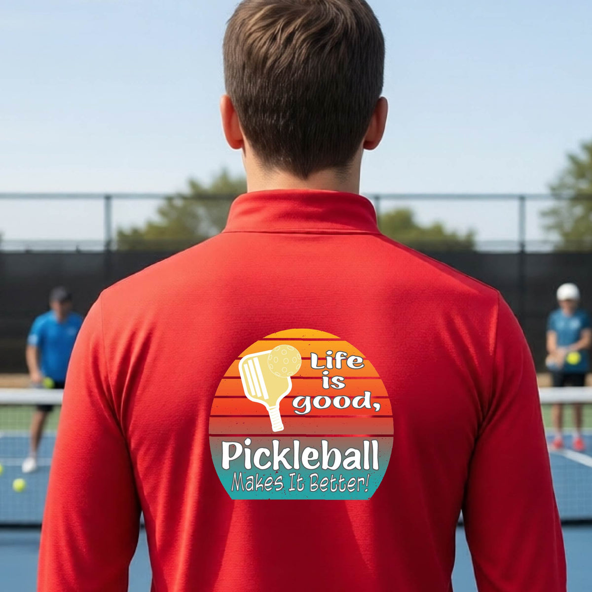 A man viewed from behind on an outdoor pickleball court, wearing a red long-sleeve quarter-zip shirt with a large colorful graphic on the back. The graphic features a retro sunset design with a pickleball paddle and the text 'Life is good, Pickleball Makes It Better!'.